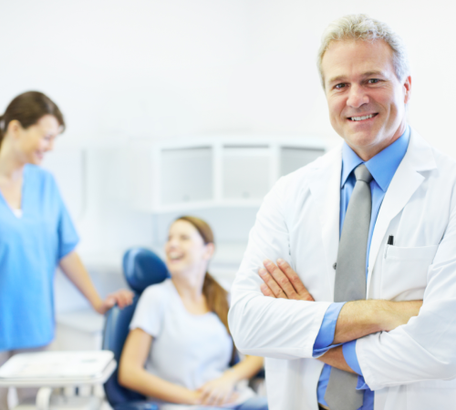 Smiling dentist with arms crossed while a patient talks with an assistant behind him